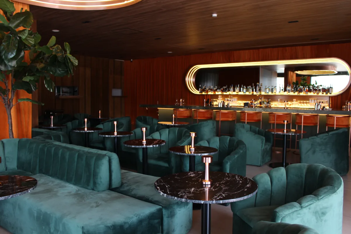 Salón de bar con sillones tapizados en verde, mesas redondas de mármol y barra iluminada al fondo