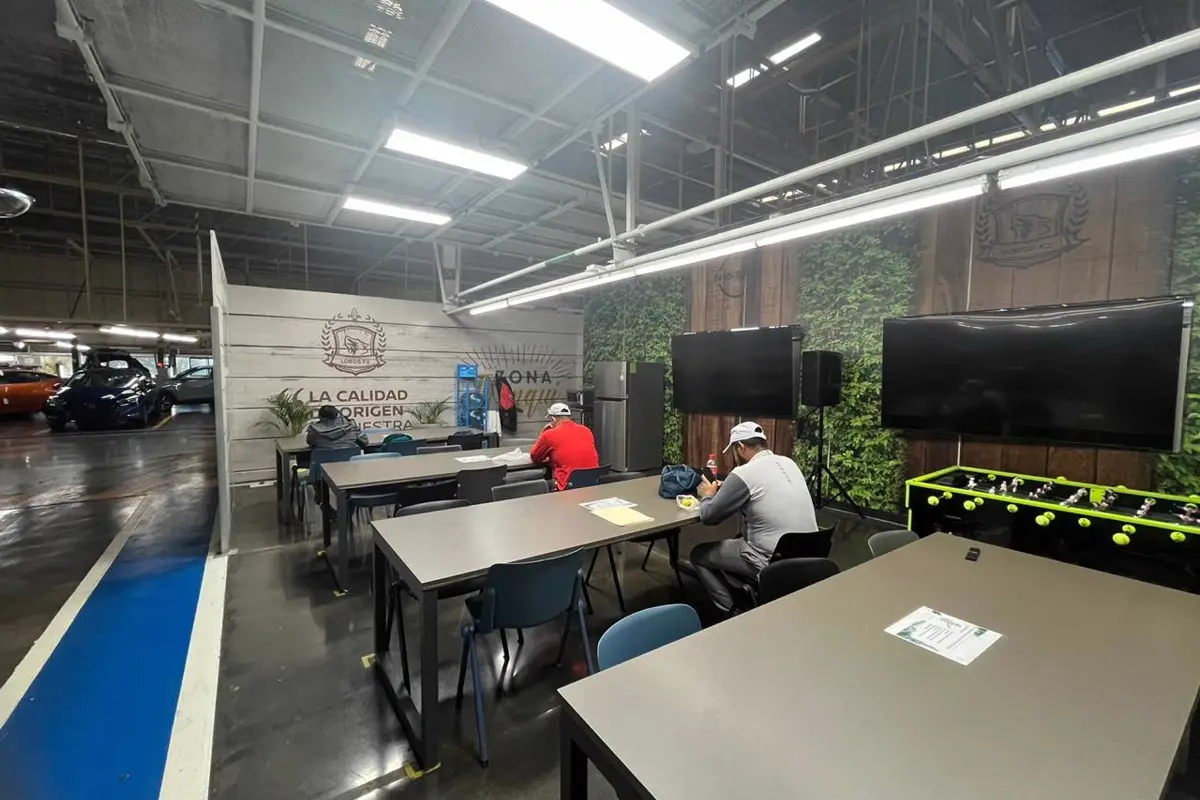 Área de comunicación en planta con mesas, sillas y pantallas sobre muro verde